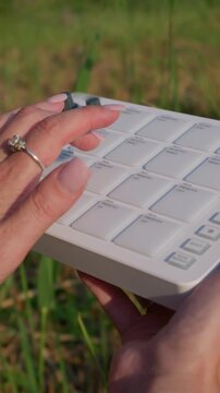 Female hands tap on a portable white MIDI drum pad controller outdoors in the grass. Close-up visual details focus on tactile rubber buttons during outdoor music production.
