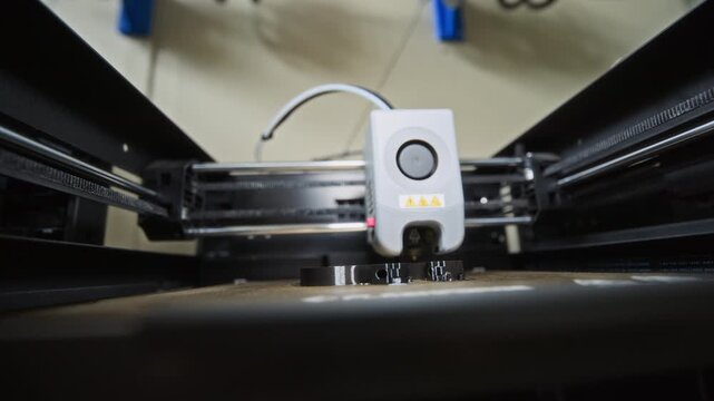 3D printer manufacturing plastic robotic component, extruding black filament layer by layer. Ideal for videos on rapid prototyping, additive manufacturing, and robotics engineering