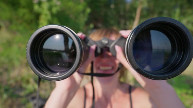 A woman looks through binoculars directly towards the viewer with her face blurred behind the optics. Concept of observation, curiosity, surveillance, and looking forward.