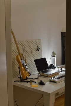 Electric guitar on a stand sits next to a home studio desk setup featuring a laptop and a MIDI keyboard
