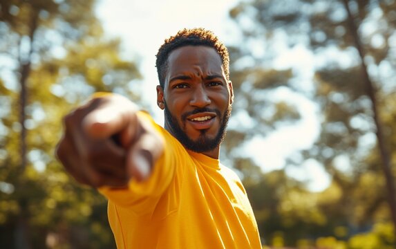 A young man in a yellow shirt pointing at the camera with a determined look on his face.  He is outdoors in a park, surrounded by greenery and sunshine.
