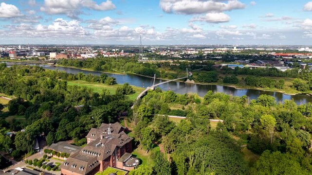 Drone view of Herrenkrug pedestrian bridge crossing Elbe River in Magdeburg, surrounded by green parkland and urban scenery. Summer cityscape shows waterfront, trees, architecture and transport links