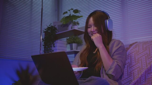 Young Asian woman focused on laptop. Eat snack There are smiles, laughter, and expressions of excitement with content on screen. Concept of modern leisure lifestyle and digital entertainment streaming