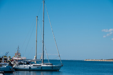 A white sailing yacht with furled sails and a dinghy secured on deck rests calmly in blue waters against a clear sky and distant coastline. © Andrey