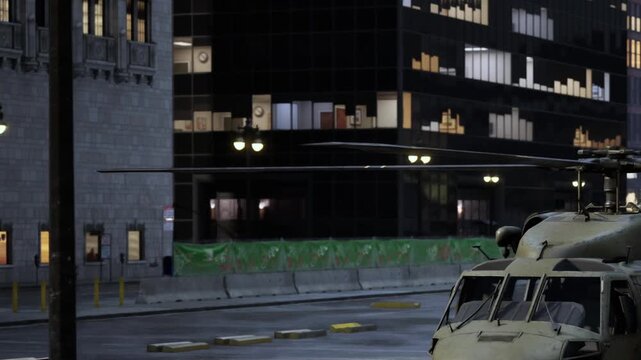 A military helicopter is parked in a street near a tall city building. Lights are on in some windows. It is night and the area is quiet. Construction barriers are visible nearby.