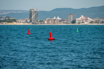 red and green navigational buoys float in the calm blue sea, with a bustling coastal resort town and a crowded sandy beach set against a mountainous backdrop in the distance. © Andrey