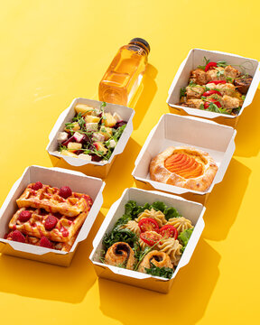 Daily healthy food delivery ration with waffles meat salad pastry and juice in containers on yellow background