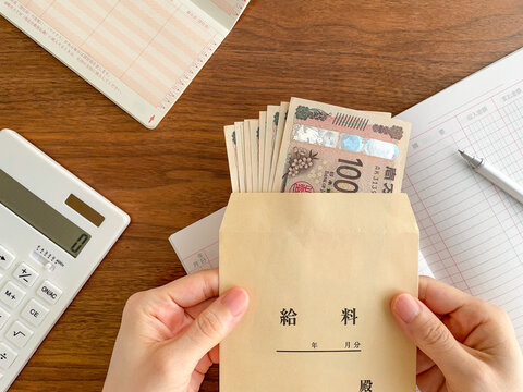 木製テーブルでお金の入った給料袋を持つ女性の手／家計管理イメージ