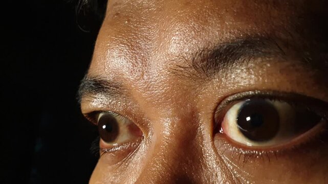 Macro close up view of an adult Indonesian Asian male eyes opening wide and looking around in dramatic low key lighting, showing glossy sclera, iris reflections, and active ocular