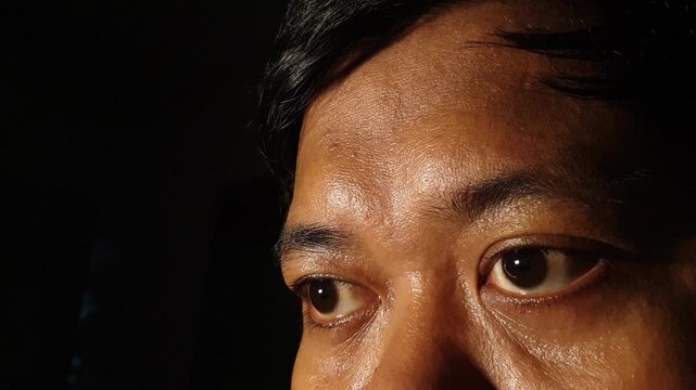 Frontal close up view of an adult Indonesian Asian male face concentrating on both eyes and forehead under dramatic low key studio light, capturing natural skin detail and steady g