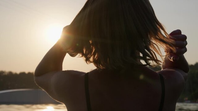 A woman gently adjusts her short bob hair in slow motion at the lake during sunset. Concept of self-care, peaceful relaxation, golden hour glow, carefree mindfulness, and wellness.