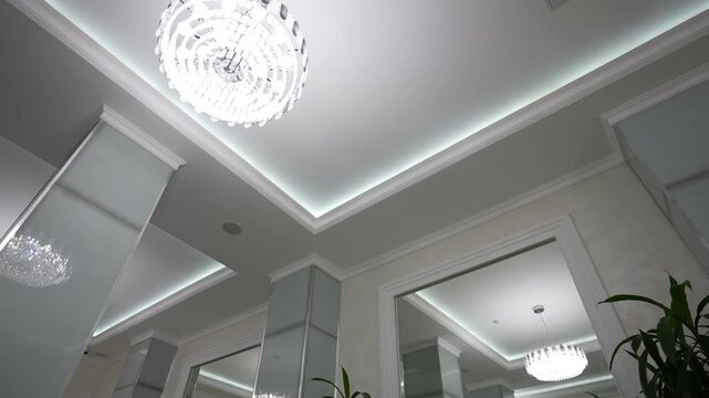 Low-angle interior tilt showing glowing crystal chandelier and recessed tray ceiling with cool LED cove lighting and mirrored reflections, with chandelier, ceiling, cove, lighting detail
