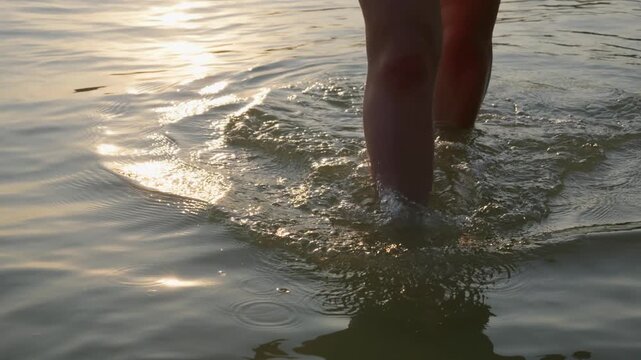 Bare legs wade through shallow, calm lake water during a peaceful sunset in slow motion. Circular ripples spread gently across the surface, reflecting bright golden evening light.