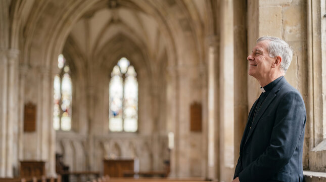 Clergyman in cassock stands inside a cathedral looking up and smiling