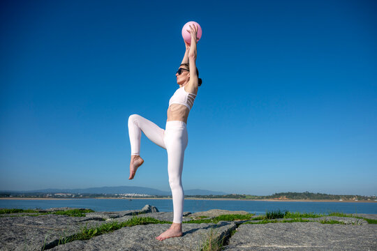 Fit woman holding an exercise ball overhead stretching outdoors, against a blue sky
