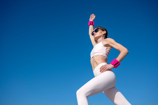 Fit woman wearing sportswear arm raised exercising outdoors with pink wrist weights against a blue sky