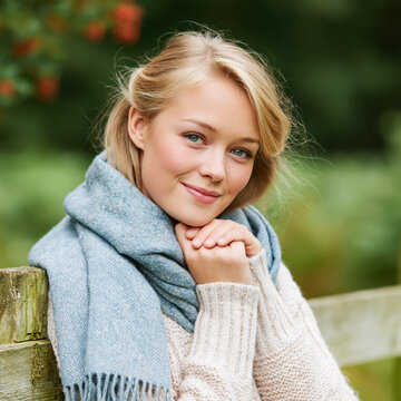 Young blonde woman with blue eyes smiling cheerfully, wearing a cozy blue scarf and an off white sweater in an autumn outdoor setting