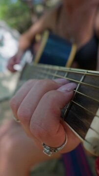 A woman plays chords on an acoustic guitar neck shown from a low angle. Shallow depth of field highlights her fingers pressing the steel strings under soft natural outdoor sunlight.