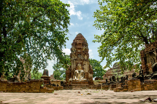 Ancient seated Buddha statue before brick prang at Wat Mahathat temple in Ayutthaya, Thailand. Framed by green trees with stone courtyard, historical ruins under blue sky. UNESCO World Heritage Site