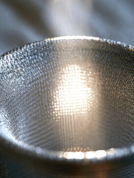 Silver mesh bowl reflecting sunlight through textured metal surface, close up study showing shimmer, curvature, selective focus, and abstract industrial form