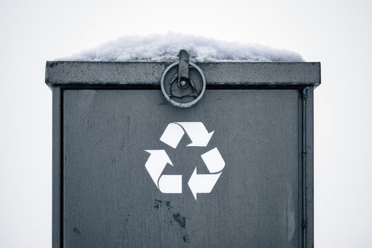 Snowy metal recycling container with white recycling symbol, viewed close up against bright winter background
