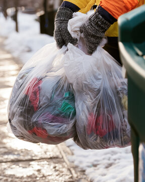 Adult hands in wool glove carrying clothing donation bag beside winter container on snowy sidewalk, civic generosity