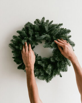Dark evergreen wreath mockup close up with hands arranging foliage on clean wall, natural light, headless framing, and blank space for holiday decor concept