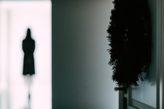 Dark evergreen wreath hanging beside minimalist doorway with blurred human silhouette bright hallway light, creating quiet seasonal scene with spacious architectural background and dramatic