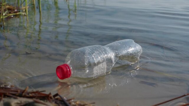 An empty clear plastic bottle with a red cap floats along the shallow sandy lake shore. Concept of environmental pollution, plastic waste crisis, ecological damage, and recycling.