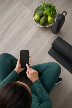 Overhead view of woman holding smartphone with blank screen on floor. Fitness tracking, diet mobile application, and healthy lifestyle concept