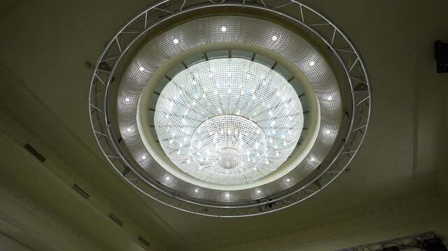 Overhead crystal chandelier illuminated within ornate ceiling and encircled by metal truss, with chandelier, crystal, lighting, ceiling detail, showing truss, diffuser, spotlight, ring context
