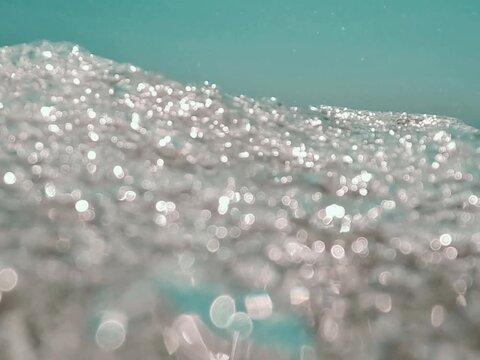 V&iacute;deo art&iacute;stico en primer plano de espuma marina con brillos, textura y desenfoque sobre agua turquesa.