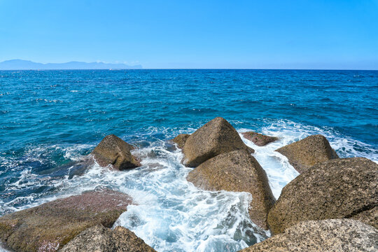Blue Mediterranean Sea waves crashing against large rocks along the rugged coast of Sicily, Italy, with distant mountains on the horizon under a bright summer sky.