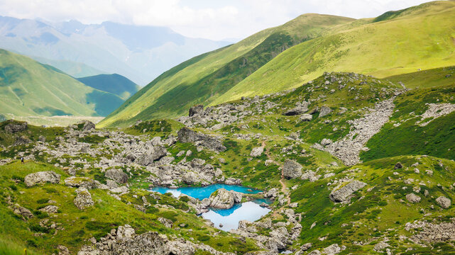 A brilliant blue mountain lake and winding trail lie among rugged rocks and green slopes in the Abudelauri Lakes area of Georgia, conveying the freedom and energy of a summer trek