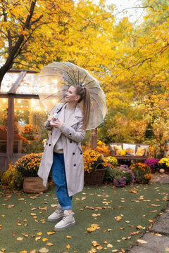 Female standing with transparent umbrella in vibrant autumn park near illuminated glasshouse. Warm rainy weather fall lifestyle concept, colorful chrysanthemums, and seasonal garden landscape.