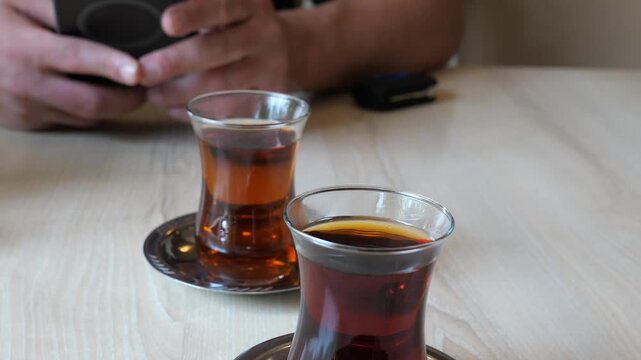 Traditional Turkish tea glasses on a wooden table with a person using a smartphone in the background