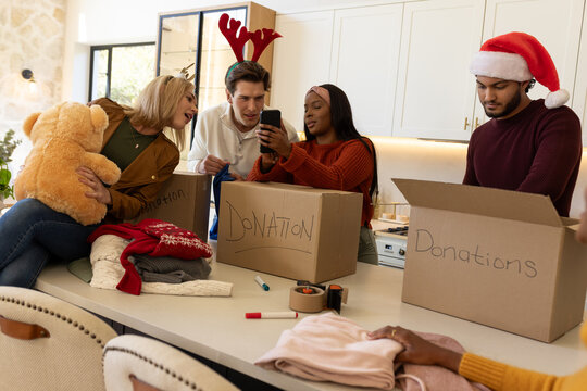 Diverse friends wearing festive hats sorting and packing donations in kitchen with teddy and phone