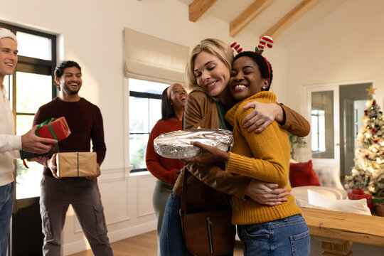 Diverse friends hugging in living room, women sharing foil dish and gifts, one wearing headband