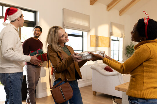 Diverse friends exchanging foil-covered dish and wrapped gifts in living room with festive hats