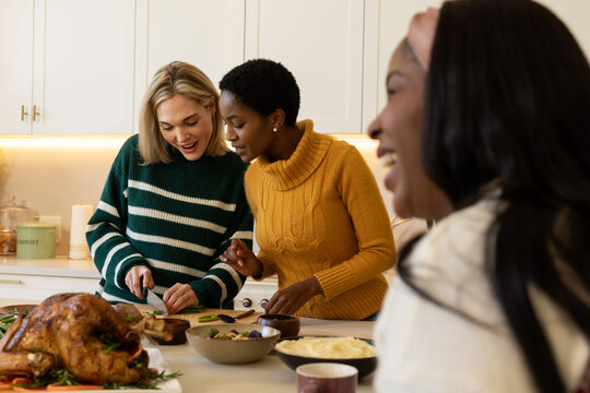 Diverse female friends wearing sweaters making meal at kitchen island arranging roasted turkey
