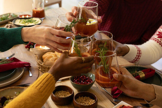 Diverse friends wearing sweaters raising stemless glasses with rosemary and citrus at wooden table