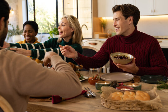 Diverse friends passing olive bowl around wooden dining table in kitchen reaching for rolls