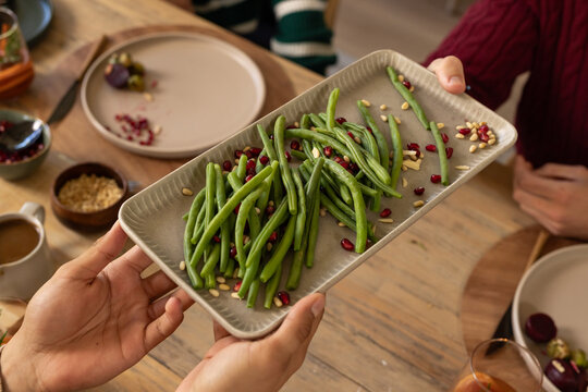 Friends passing ceramic platter with green beans, pomegranate arils and pine nuts at dining table