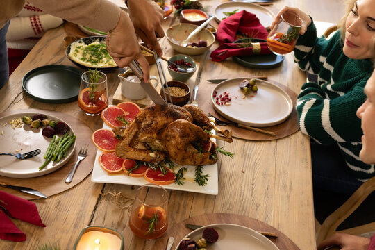 Diverse family carving roasted whole turkey at home using carving knife while holding amber glasses