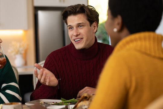 Diverse friends sitting at kitchen table, maroon sweater man speaking, plates and glass visible