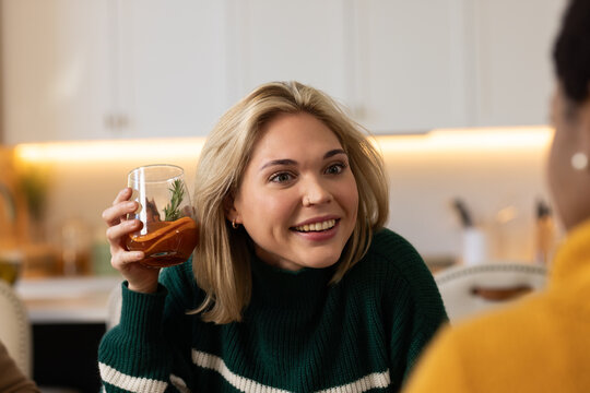 Diverse female friends chatting at kitchen table, blonde in green sweater holding tumbler with herb