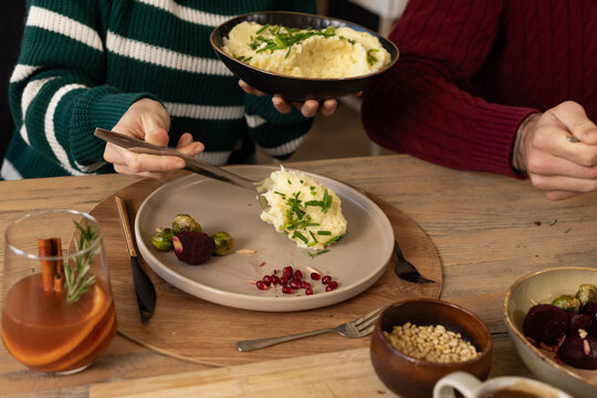 Couple in striped and maroon sweaters serving mashed potatoes from black bowl at wooden table