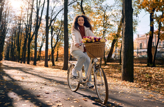 Happy smiling young woman riding a bike with a basket of flowers in a golden autumn park. Cozy slow morning weekend lifestyle.