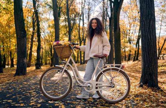 Smiling young woman with curly hair standing beside a white classic city bicycle with a wicker basket full of colorful flowers on a scenic pathway in a beautiful autumn park.