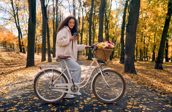 Beautiful woman standing in an autumn park with a white vintage bicycle featuring a wicker basket filled with flowers,looking at her smartphone. Concept of recreation, and enjoying the autumn season.